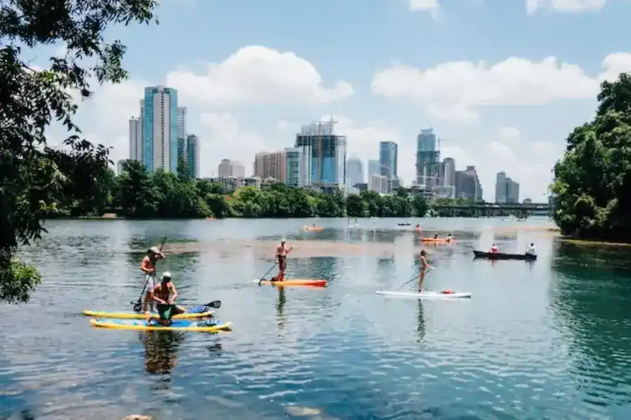 A Landscape in Austin showing many small boat riders on a lake with many trees behind them, followed by skyscrapers